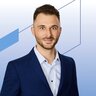 Ein Mann mit Bart, der einen marineblauen Blazer und ein hellblaues Hemd mit Knöpfen trägt.