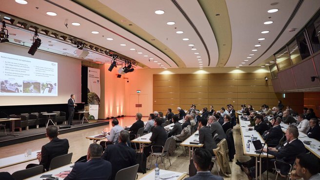 People sit in a large conference room with a presentation screen in the background.