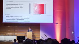 A man is presenting behind the podium of an atmospherically lit stage. A slide on the subject of enhanced fast charging can be seen on the screen.