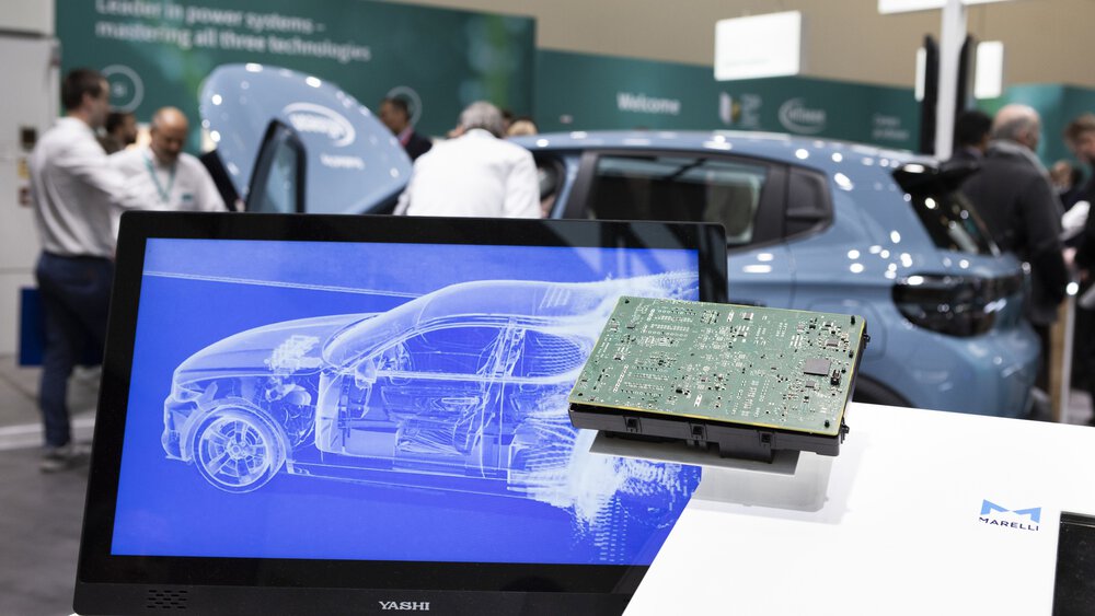 An einem Messestand ist auf einem Monitor ein stilisiertes Auto zu sehen. Daneben liegt einer Leiterplatte.