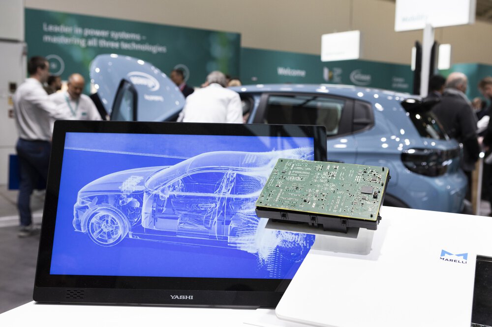 An einem Messestand ist auf einem Monitor ein stilisiertes Auto zu sehen. Daneben liegt einer Leiterplatte.