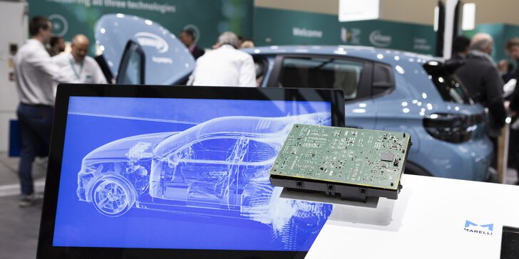 An einem Messestand ist auf einem Monitor ein stilisiertes Auto zu sehen. Daneben liegt einer Leiterplatte.