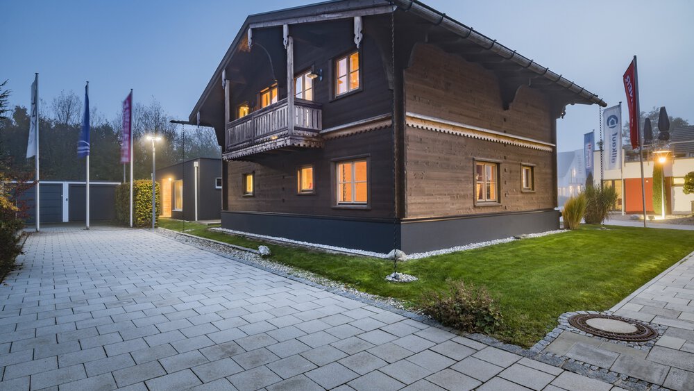 Modernes zweistöckiges Haus mit großen Fenstern, umgeben von einem Rasenhof und einer gepflasterten Auffahrt in der Abenddämmerung.