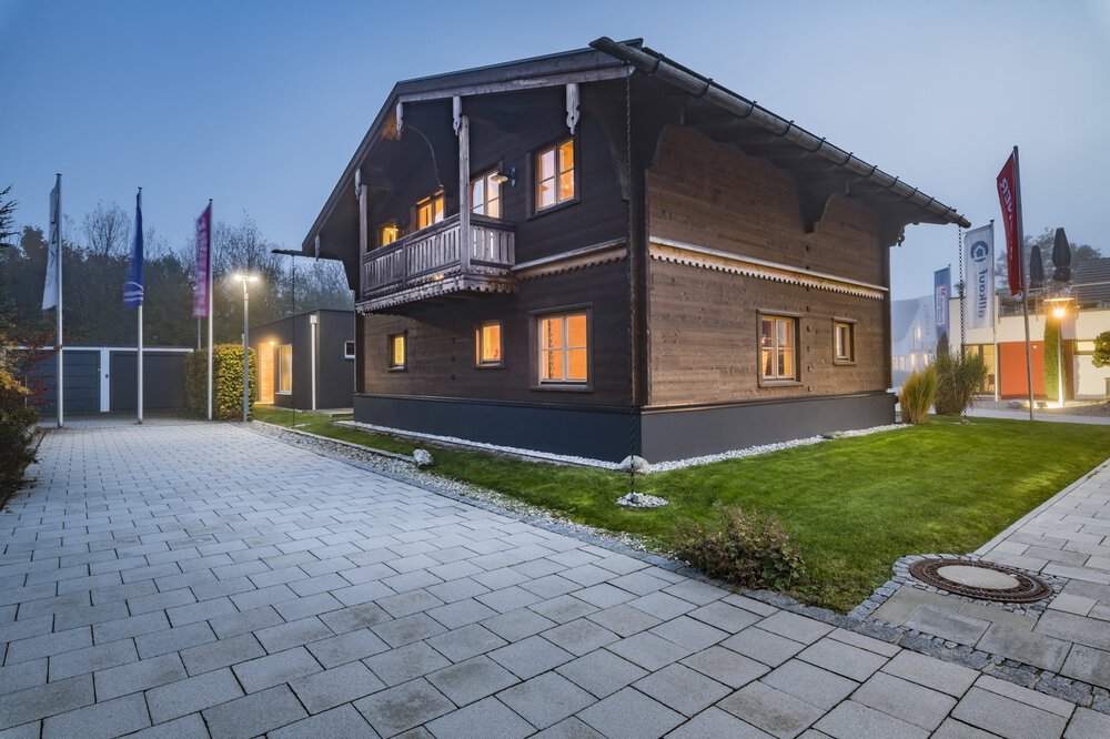 Modernes zweistöckiges Haus mit großen Fenstern, umgeben von einem Rasenhof und einer gepflasterten Auffahrt in der Abenddämmerung.