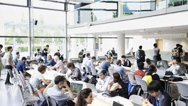 A bright, modern workspace where people sit at long tables in front of their laptops while others walk past.