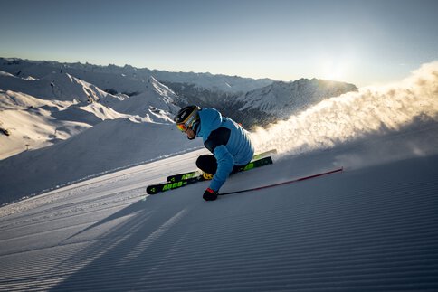 Ein Skifahrer in blauer Jacke und Helm carvt auf Fischer Curv GT-Skiern einen verschneiten Hang hinunter und hinterlässt eine Spur aus Pulverschnee vor der Kulisse der sonnenbeschienenen Berge.