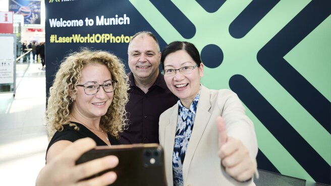 Drei Teilnehmende machen ein Selfie vor dem Welcome-Board der Laser World of Photonics 2025 in München.