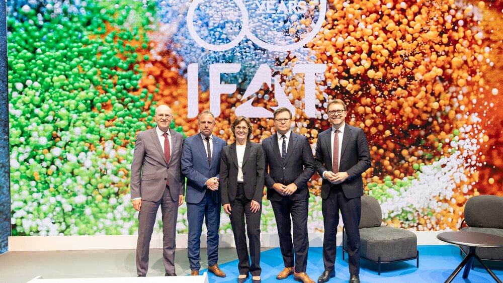Five people in business attire posing on a stage in front of a large LED wall displaying the 