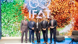 Five people in business attire posing on a stage in front of a large LED wall displaying the 