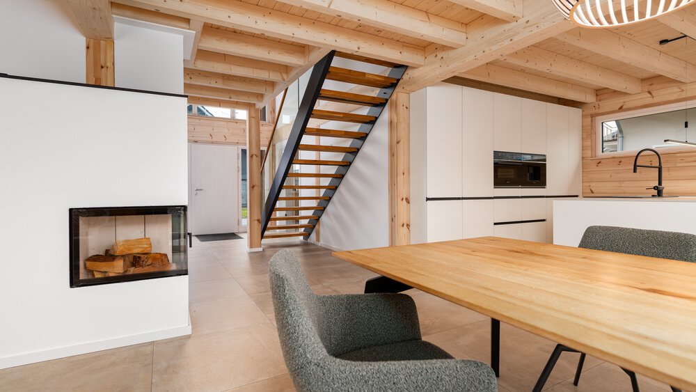 Moderner heller Essbereich mit Holztisch, Stühlen und Kamin und Blick auf den Eingangsbereich mit Holztreppe zum Obergeschoss.