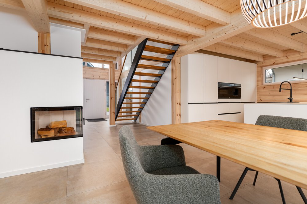 Moderner heller Essbereich mit Holztisch, Stühlen und Kamin und Blick auf den Eingangsbereich mit Holztreppe zum Obergeschoss.