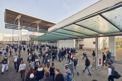 Eine Menschenmenge bewegt sich auf den Eingang der Messe München unter einem überdachten Weg zu.