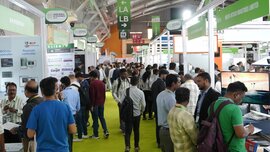A look down one of the aisles at Laser World of Photonics India. Numerous visitors walk across a bright green carpet and view the exhibition stands.