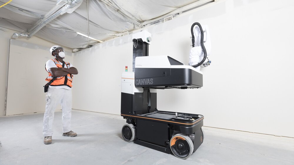 Eine Person in Sicherheitsweste beobachtet einen großen, fahrbaren Maschinenroboter in einer Indoor-Baustellenszene.