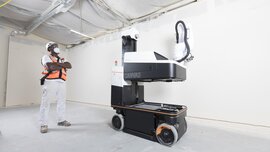 Eine Person in Sicherheitsweste beobachtet einen großen, fahrbaren Maschinenroboter in einer Indoor-Baustellenszene.