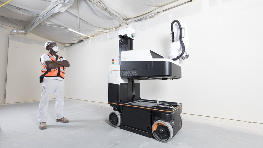 A person in a safety waistcoat observes a large, mobile machine robot in an indoor construction site scene.