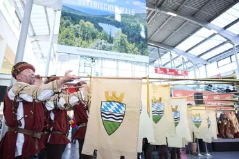 People with brass instruments and the flag of the Bavarian Forest perform in a trade fair entrance hall.