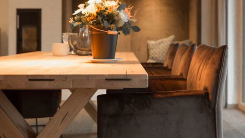 Ein hölzerner Esstisch mit Blumenarrangement, umgeben von gepolsterten Stühlen, in einem gemütlichen, warm beleuchteten Raum.