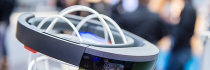 A sleek, futuristic AR headset with a transparent visor, displayed on a white wire head model at a trade fair stand.