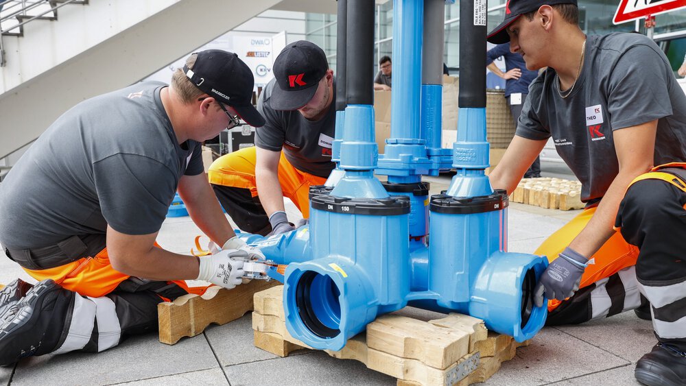 Three workers in protective equipment load a large blue part of a piping system in front of an exhibition hall.