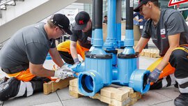 Three workers in protective equipment load a large blue part of a piping system in front of an exhibition hall.