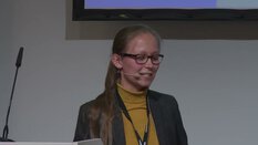 Frau mit Brille und langen Haaren, gelbem Oberteil und grauer Jacke spricht mit Headset an einem Rednerpult.