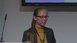 Woman with glasses and long hair, yellow top and grey jacket speaks with headset at a lectern.