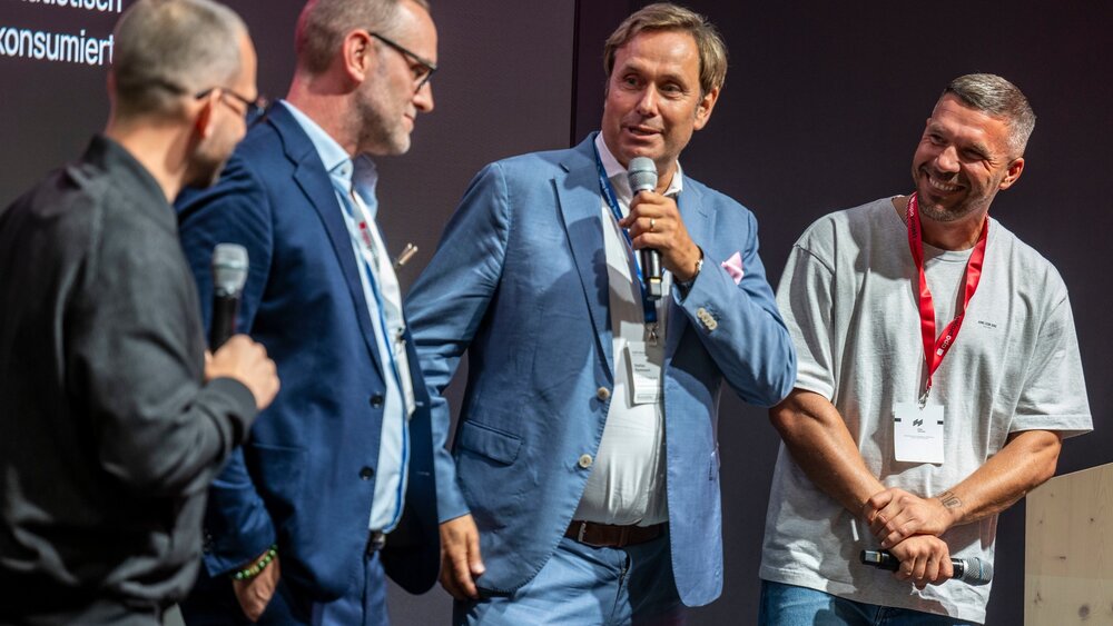 Four men on stage at EXPO REAL 2026, two holding microphones, engaged in lively discussion and smiling before a dark event backdrop.