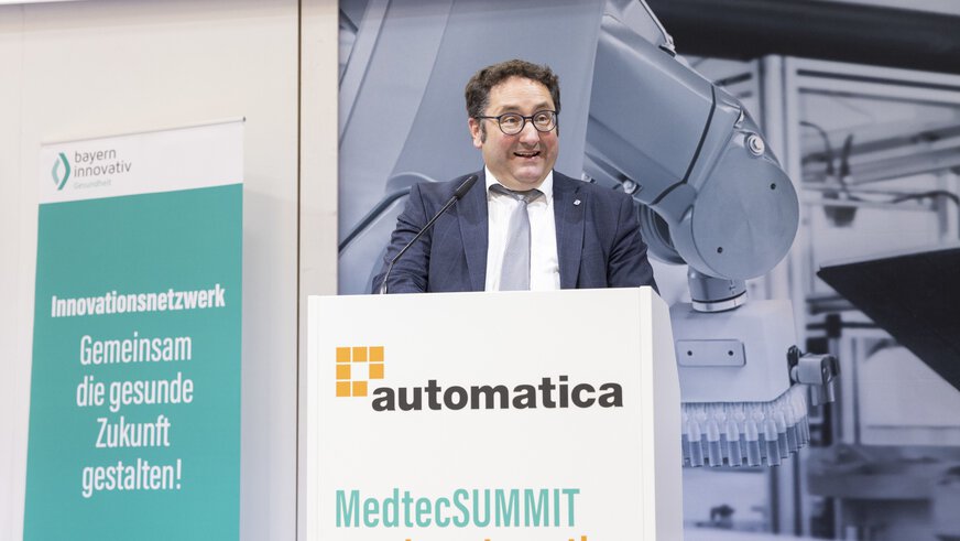 Ein Redner im Anzug steht am Rednerpult der „automatica“, das mit dem Schriftzug „MedtecSUMMIT meets automatica“ beschriftet ist. Im Hintergrund sind ein Roboterarm und ein Roll-up des Netzwerks „bayern innovativ“ mit dem Slogan „Gemeinsam die gesunde Zukunft gestalten!“