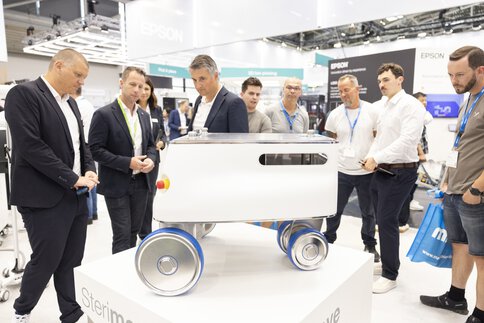 Group of men viewing white wheeled robot at trade fair presentation.