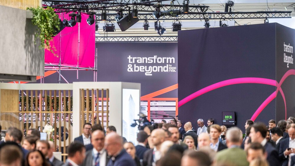 Eine große Menschenmenge nimmt an einer Business-Veranstaltung in einem Saal teil, im Hintergrund sind ein „Transform & Beyond“-Schild und farbenfrohe Ausstellungsstücke zu sehen. Die Teilnehmer knüpfen Kontakte und unterhalten sich.