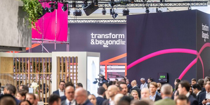 Eine große Menschenmenge nimmt an einer Business-Veranstaltung in einem Saal teil, im Hintergrund sind ein „Transform & Beyond“-Schild und farbenfrohe Ausstellungsstücke zu sehen. Die Teilnehmer knüpfen Kontakte und unterhalten sich.