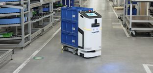 A white and black transport robot labelled ‘rexroth’ transports three blue crates in a warehouse with many full shelves.