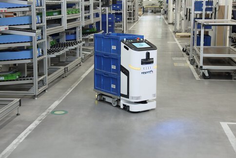 A white and black transport robot labelled ‘rexroth’ transports three blue crates in a warehouse with many full shelves.