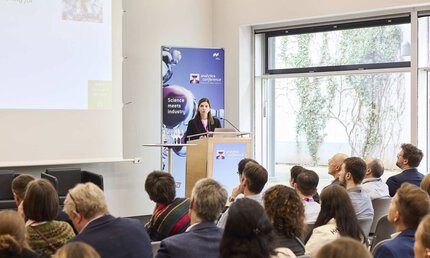 Eine langhaarige Person hält einen Vortrag. Mehrere Personen hören interessiert zu.