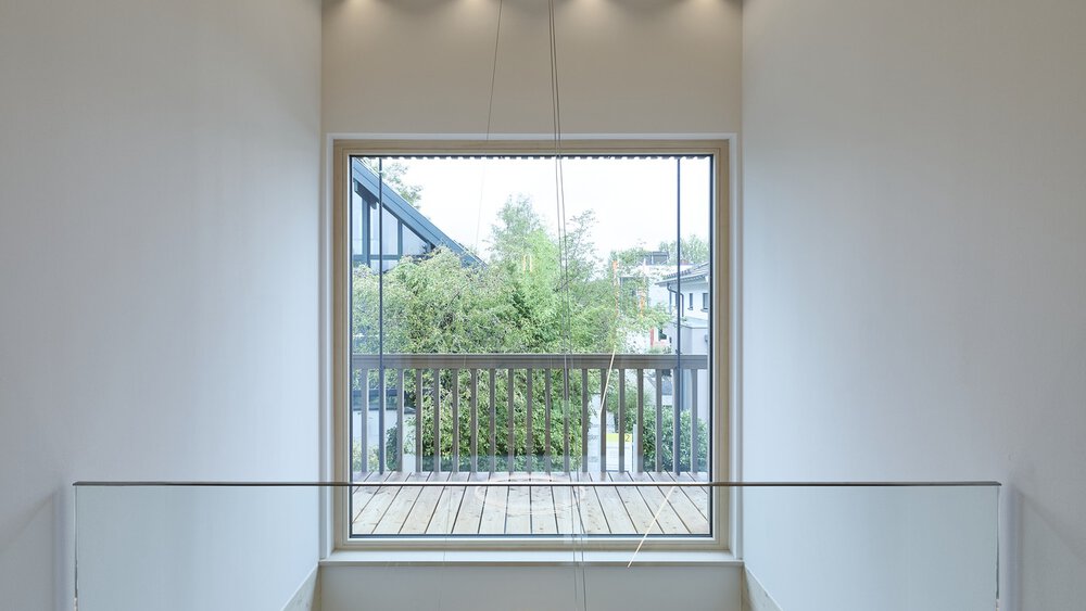 Großes Fenster mit Blick auf einen Balkon mit grünem Laub, eingerahmt von minimalistischen weißen Wänden und einer Holzlattendecke.