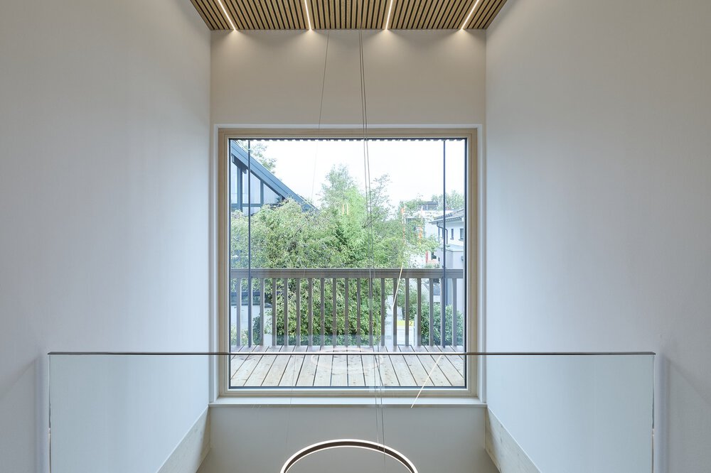 Großes Fenster mit Blick auf einen Balkon mit grünem Laub, eingerahmt von minimalistischen weißen Wänden und einer Holzlattendecke.