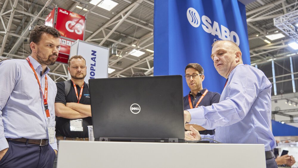 Four men stand around a laptop at a trade fair stand and discuss.
