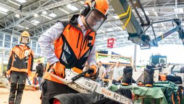 Eine Person in orange-schwarzer Schutzkleidung durchtrennt mit einer Motorsäge einen aufgebockten Baumstamm.