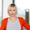 A woman with short blonde hair, a gray top, and an orange cardigan is standing in a house and looking at the camera.