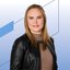 Portrait of a woman with shoulder‑length blonde hair wearing a black leather jacket and brown turtleneck sweater with a necklace in front of a light background with blue graphic lines