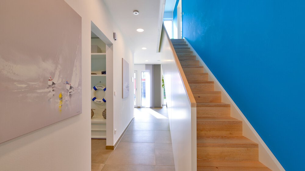 Heller Flur mit Holztreppe, weißen Wänden, blauer Akzentwand und Sonnenlicht, das durch eine Tür hereinströmt.