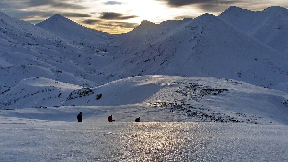 Vier Personen wandern durch eine verschneite Berglandschaft unter einem dramatischen, bewölkten Himmel bei Sonnenuntergang.
