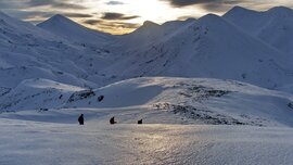 Vier Personen wandern durch eine verschneite Berglandschaft unter einem dramatischen, bewölkten Himmel bei Sonnenuntergang.