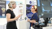 Zwei Frauen im Gespräch an einem Messestand der World of Quantum in München, mit Exponat und Analyse-Diagrammen im Hintergrund.