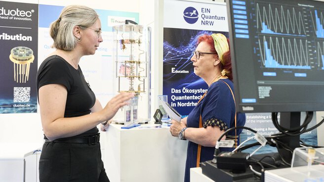 Zwei Frauen im Gespräch an einem Messestand der World of Quantum in München, mit Exponat und Analyse-Diagrammen im Hintergrund.