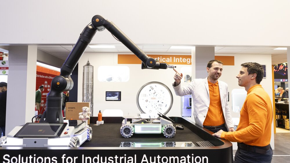 Two men in orange shirts stand next to a platform on which various industrial robots are displayed.