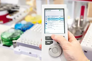 Handheld device with a screen displaying a list of items. The background is blurred and shows a laboratory environment.