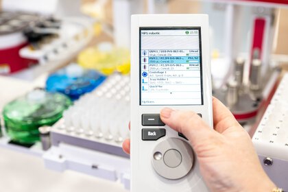 Handheld device with a screen displaying a list of items. The background is blurred and shows a laboratory environment.