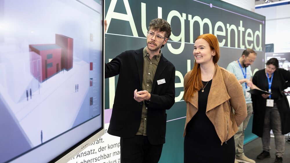 Zwei Personen stehen rechts neben einem großen Bildschirm und blicken gemeinsam auf darauf. Auf dem Bildschirm ist das Model eines Hauses zu sehen.
